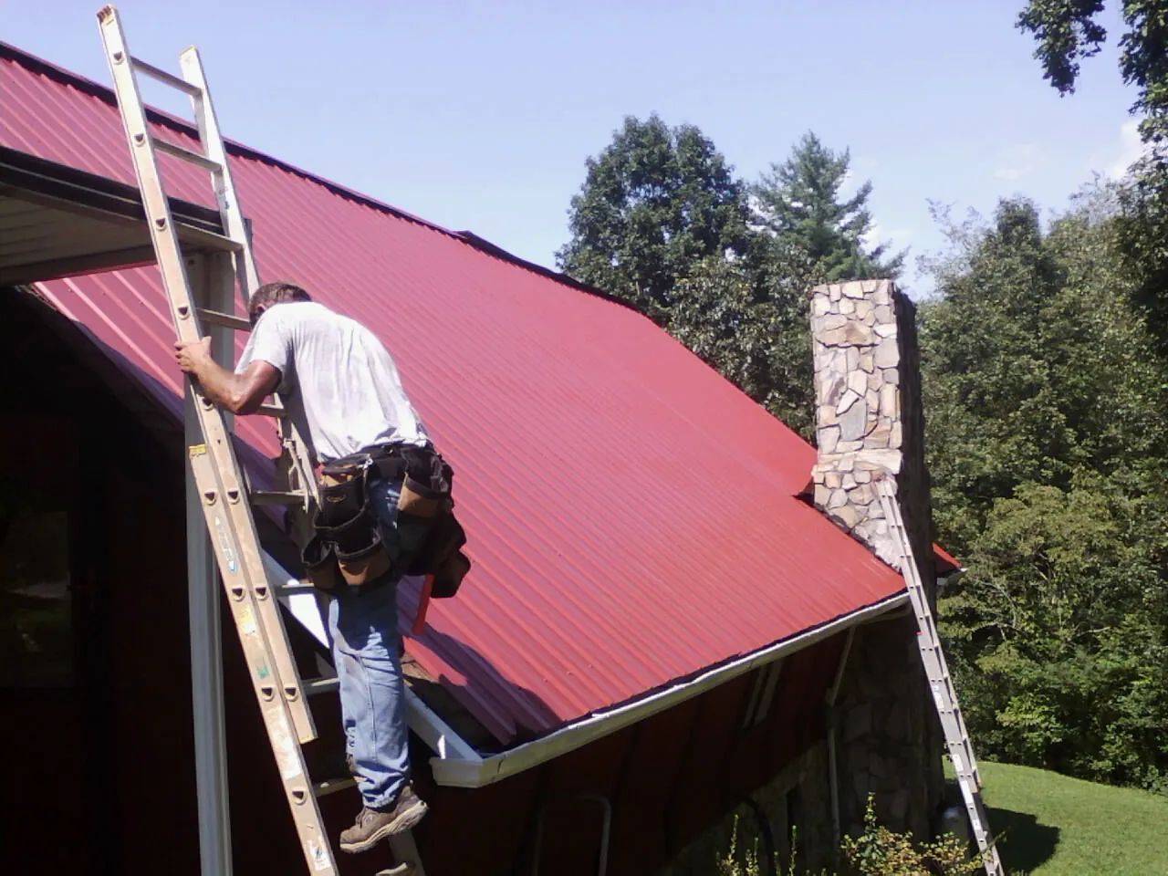 Roofer on a ladder beside a red metal roof and stone chimney — Soffit & Fascia Repair by Roofing Brothers in North Attleboro